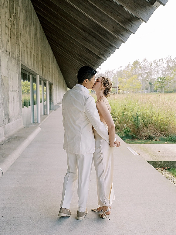 Wedding kiss portrait of bride and groom kissing in side profile, bride in satin slip dress with braided updo under modern walkway windows