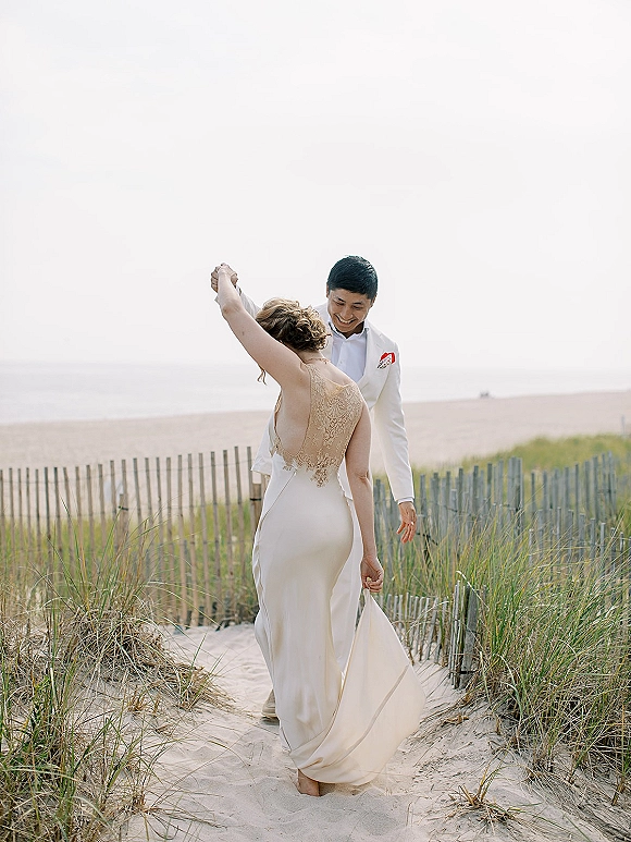 Couple portrait of a beach wedding couple twirling on a sandy dune path by ocean and sky, bride in lace dress back and groom suit