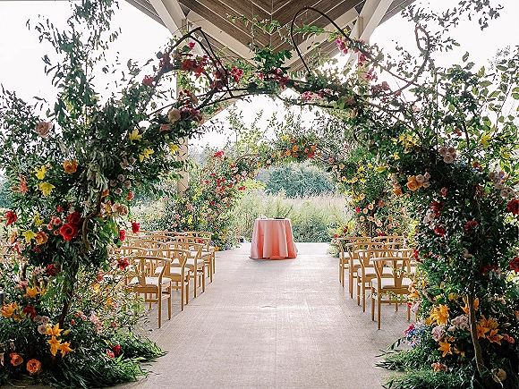 Ceremony setup with a wedding floral arch featuring hanging flowers and greenery garlands under a covered pavilion facing open fields