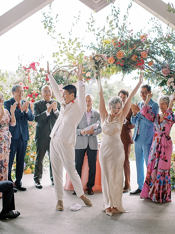 Ceremony recessional as bride and groom cheer down the aisle, bride with bouquet and birdcage veil, guests applauding under floral arch.