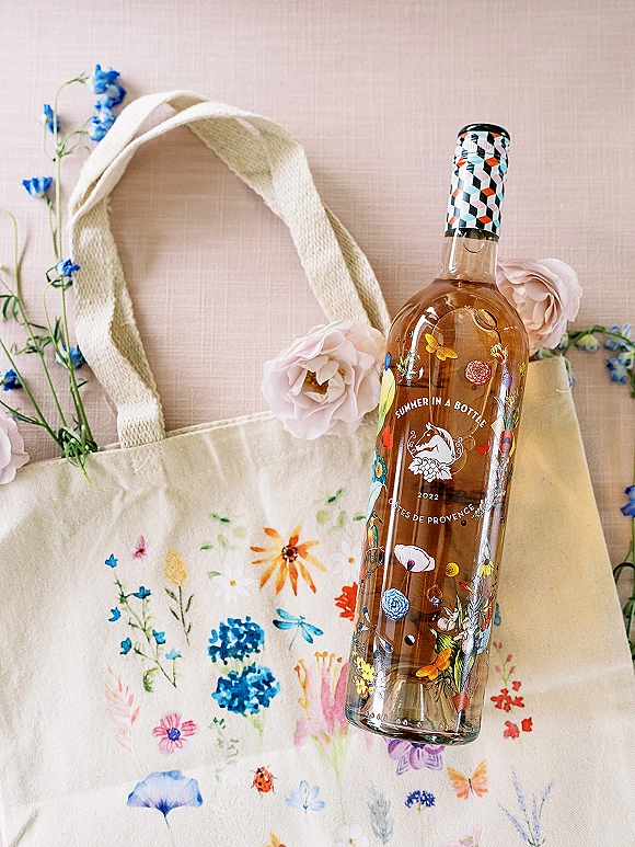 Wine bottle gift in a wedding welcome bag, showing a floral label nestled in a canvas tote with pink and blue flowers on a pink backdrop