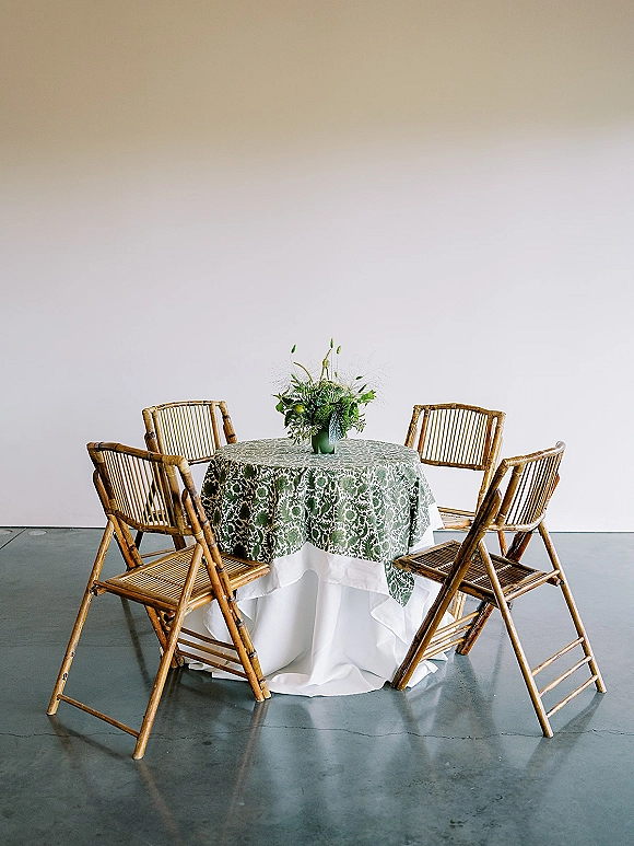 Reception tablescape with a round wedding table setup, green patterned tablecloth layered with white linen and a floral centerpiece in green vase.