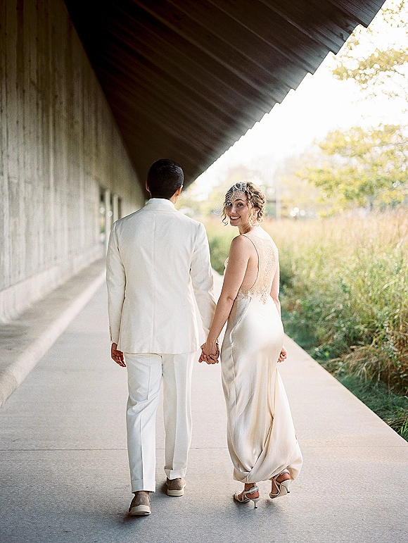 Couple portrait of bride and groom walking away holding hands, groom in a white suit and bride in a satin dress with birdcage veil, by a modern building walkway