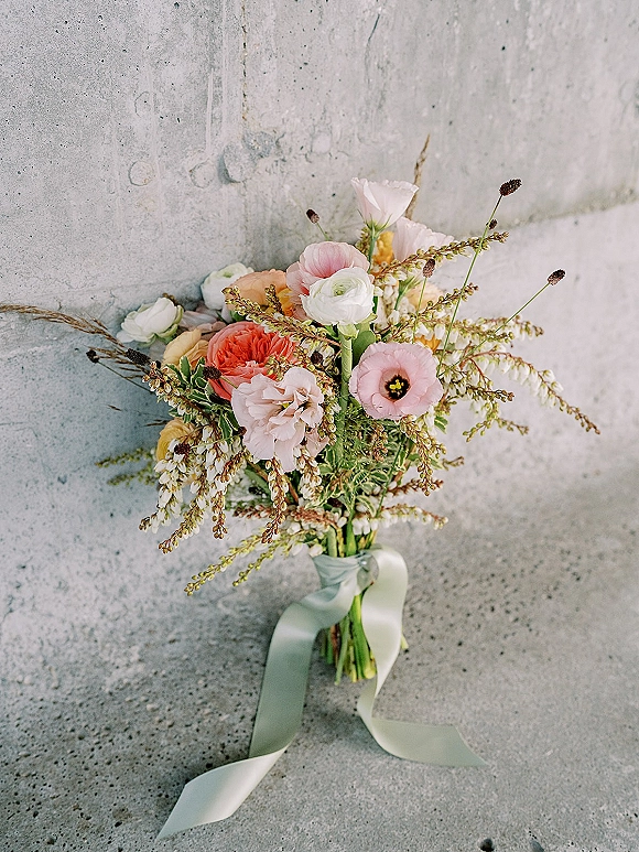 Bridal bouquet with pastel wedding bouquet blooms—ranunculus, poppy anemone, greenery, and seed pods—resting on a concrete floor with ribbon wrap