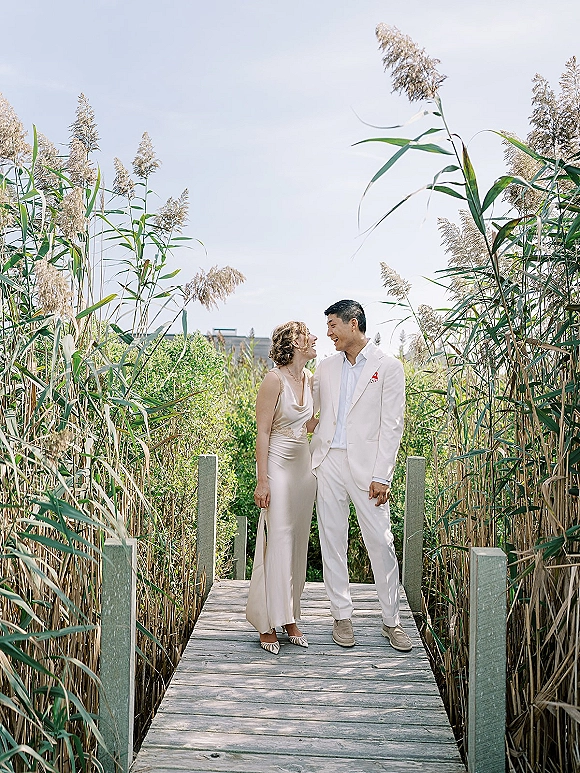 Couple portrait of bride and groom laughing while walking a wooden boardwalk through tall reeds, bride in satin slip wedding dress