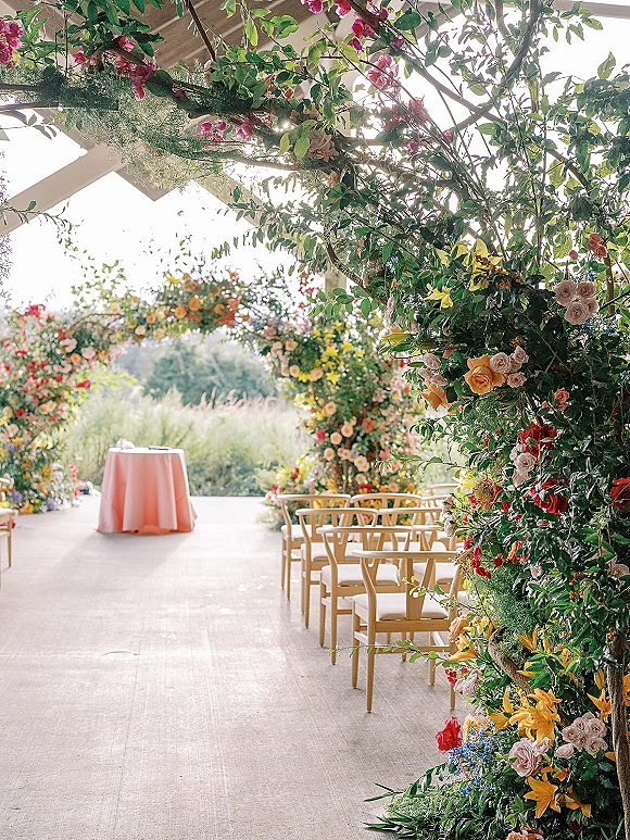 Ceremony setup with outdoor ceremony chairs under a wooden pergola, featuring a colorful floral arch and sunlit patio setting
