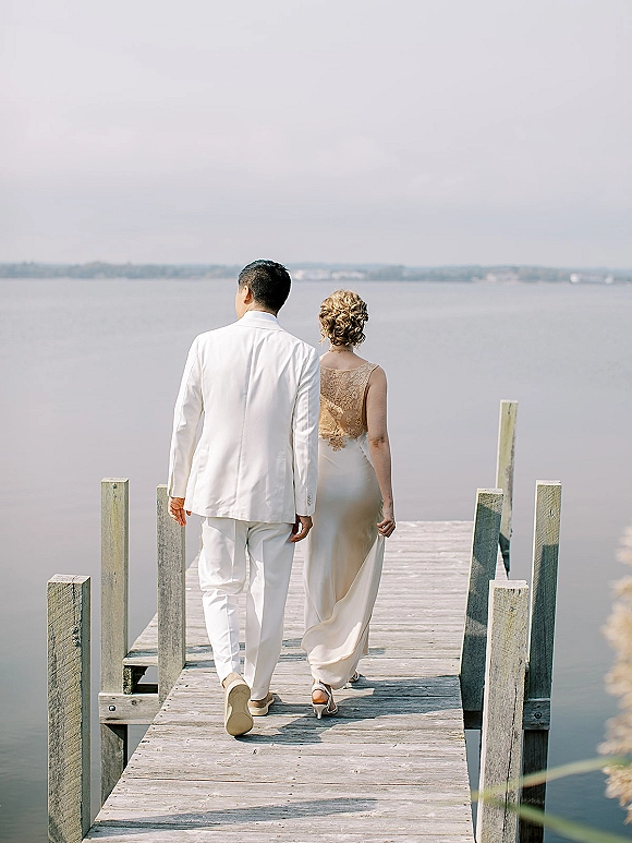 Couple portrait of newlyweds walking away on a wood dock by the lake, bride in lace bodice dress and heels beside groom in suit