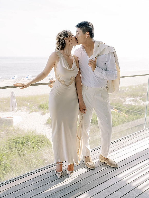 Wedding kiss portrait of bride and groom kissing on a balcony railing, ocean and sand dunes behind, bride in slip dress and pearls