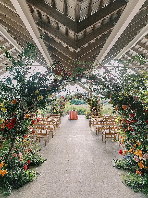 Ceremony setup with an outdoor ceremony arch of colorful flowers and lush greenery under a wood-beam pavilion overlooking field and trees