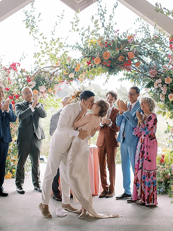 Wedding kiss under a colorful floral arch as guests applaud on a daylight outdoor patio beneath a pavilion roof, bride in satin gown and groom in suit