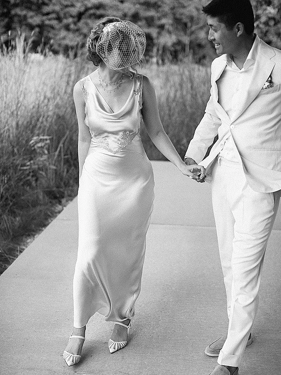 Couple portrait of bride and groom holding hands on an outdoor walkway, her birdcage veil and lace dress detail beside his white suit