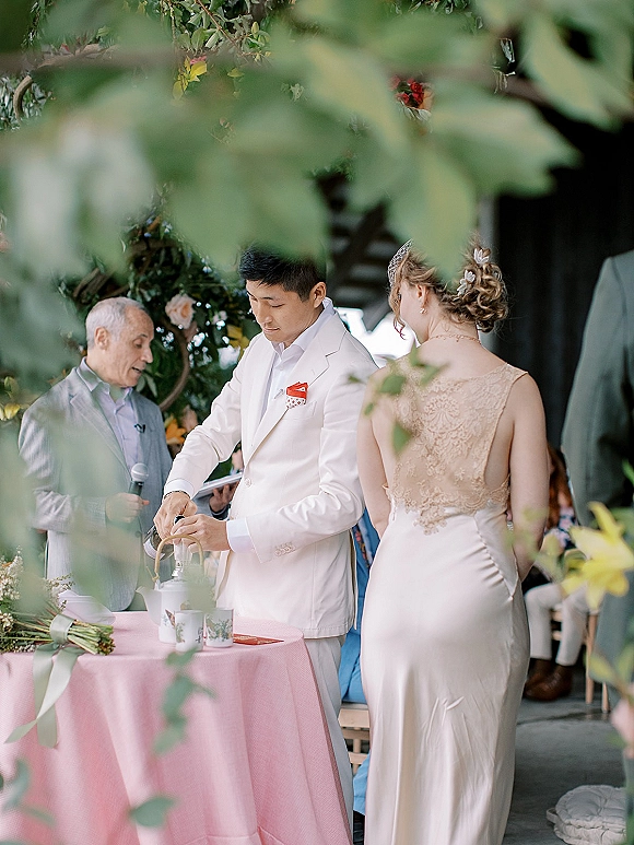 Ceremony ritual at a pink table as officiant speaks into a microphone, bride in lace-back dress and groom in white suit amid greenery