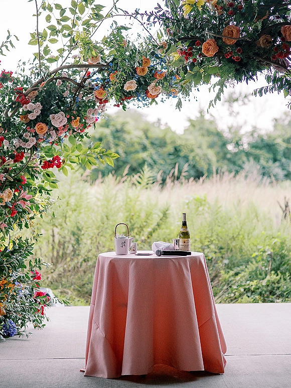 Ceremony signing table with marriage license papers, pen, microphone, and wine under a floral arch with garden roses on a patio meadow backdrop