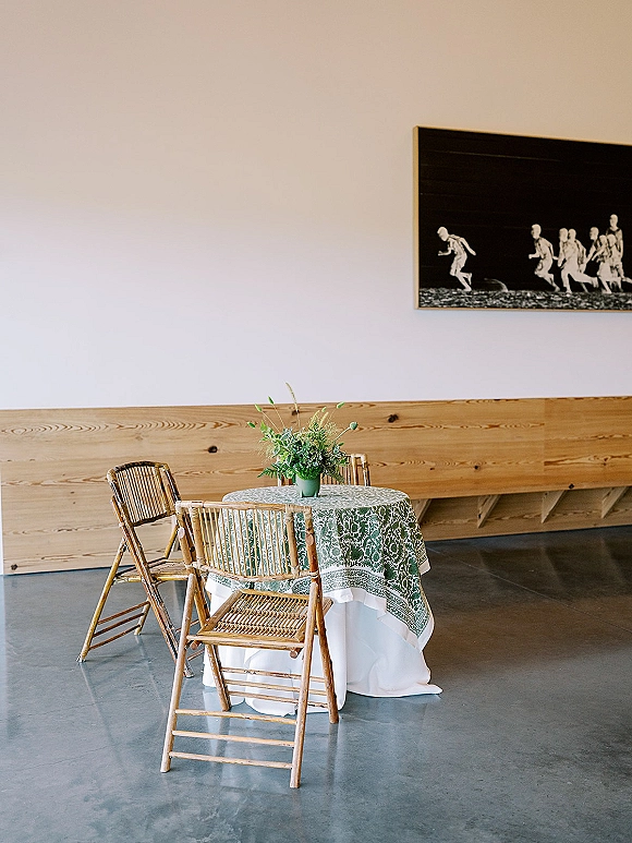 Reception table setup with a green patterned tablecloth, layered white linens, and a ceramic vase floral centerpiece in a modern room
