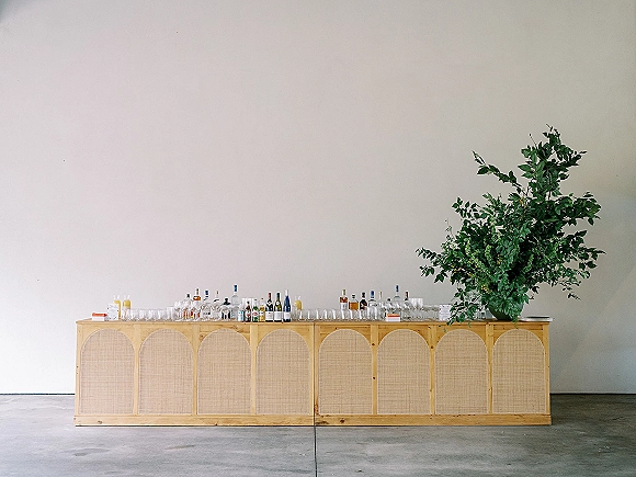Wedding bar setup with a wood bar lined with liquor and wine bottles, glassware, and greenery against a white wall on concrete floor