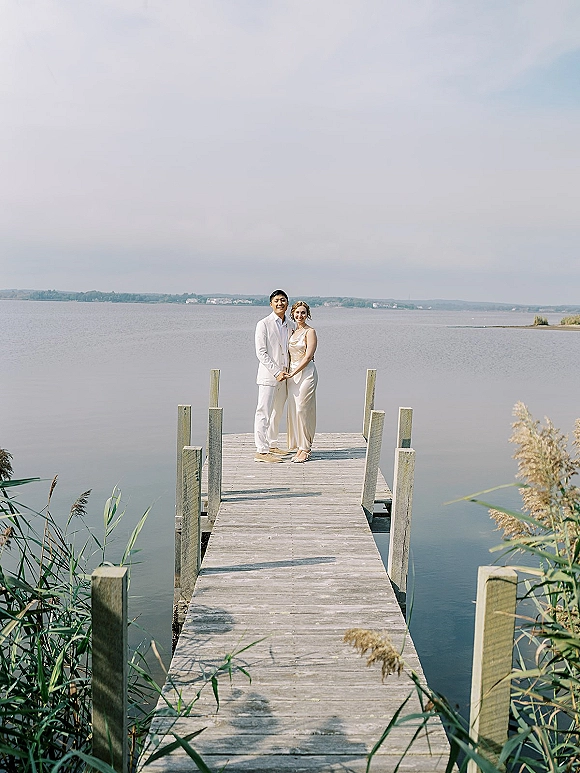 Couple portrait holding hands on a wooden dock by a lake, bride in a minimal wedding dress and groom in a white suit and sandals