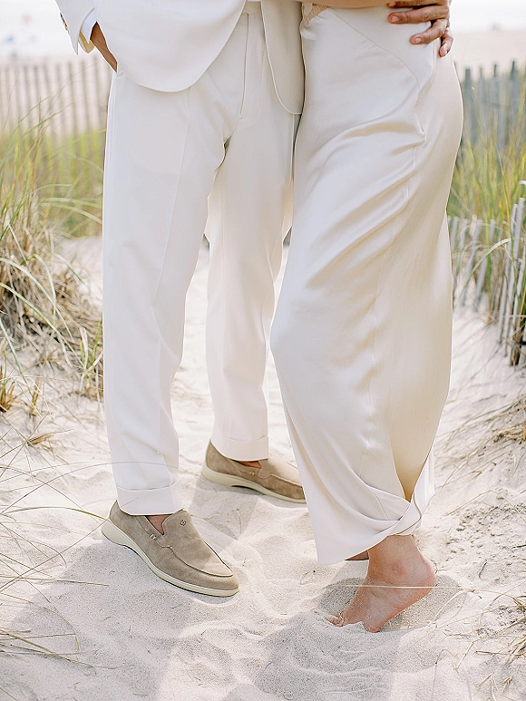 Couple portrait at a sand beach wedding, groom in white suit embracing bride in wide-leg pants, dune grass and wooden fence behind