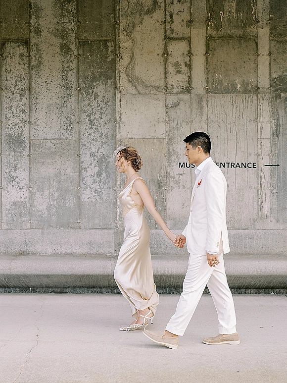 Couple portrait of bride and groom walking hand in hand in side profile, groom in white suit, bride in satin slip dress by concrete wall signage