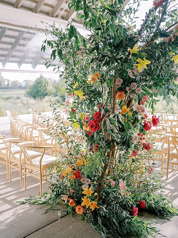 Wedding ceremony floral arch with an asymmetrical floral arch design of lush greenery and roses, lilies, and branches in an outdoor pavilion
