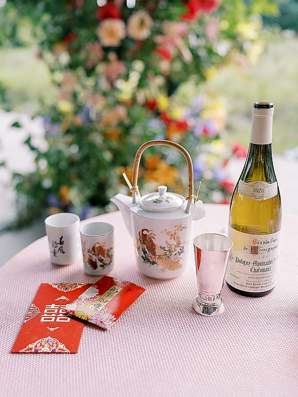 Wedding tea ceremony table with a Chinese wedding tea set, porcelain teapot and cups beside red envelopes and a wine bottle, floral bokeh behind