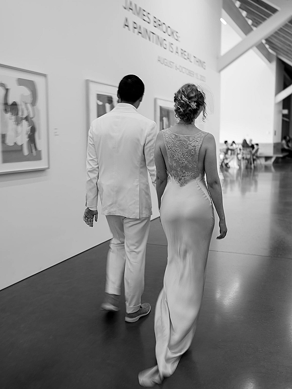 Couple portrait of bride and groom walking away holding hands, bride in lace-back gown and updo, in a modern art gallery