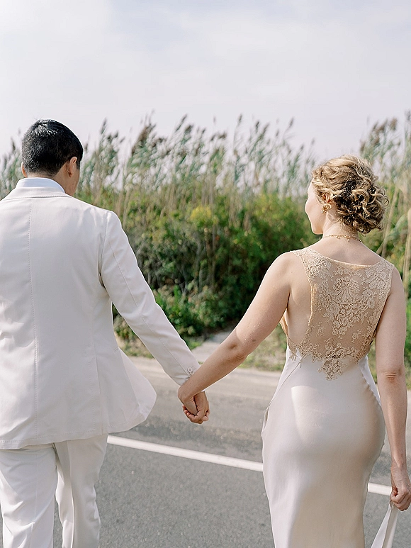 Couple portrait of bride and groom walking away holding hands, her lace-back dress and his white suit on a coastal road by beach grass