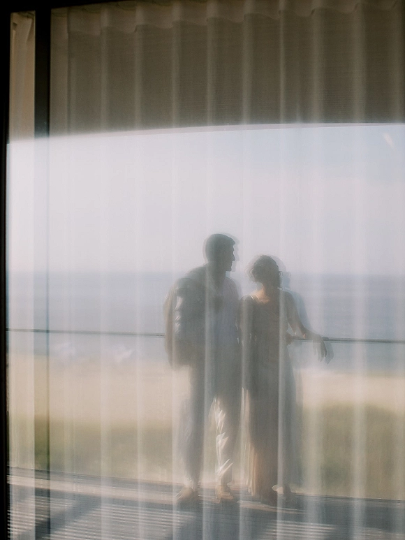 Couple portrait of bride in wedding dress and groom in suit on a balcony, framed by sheer curtains with ocean horizon beyond