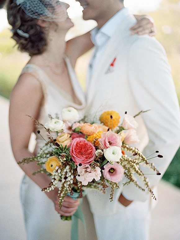 Wedding couple portrait of bride and groom embracing, holding a colorful bridal bouquet with ribbon in warm sunlight and soft greenery background