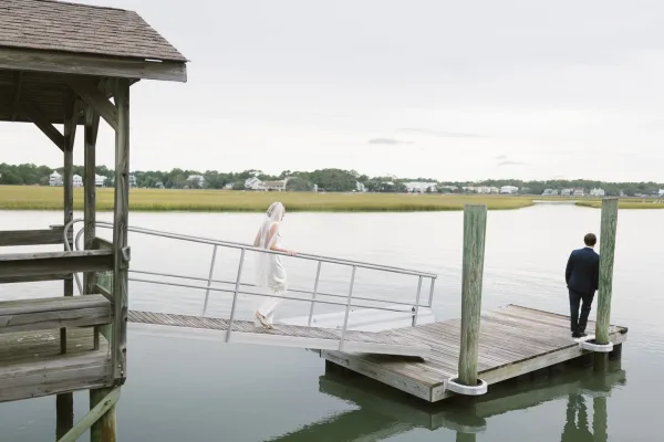 First look moment on a wooden dock as the bride in a long veil approaches the groom in a navy suit by marshy water under clouds
