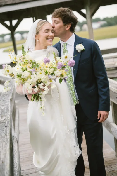 Wedding couple portrait with bride holding a wildflower bouquet as the groom gives a forehead kiss on a wooden boardwalk by the water