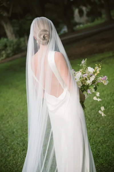 Bridal portrait of a bride from behind in a simple white dress with a long wedding veil, low bun, pearl earrings, and bouquet on a garden walkway