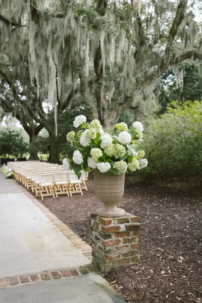 Ceremony aisle decor with wedding aisle flowers in a white hydrangea urn on a brick pedestal beside wood chairs under oak trees with Spanish moss