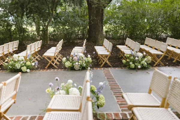 Ceremony aisle decor with hydrangeas and white daisies lining a brick and concrete aisle, woven folding chairs in a tree-shaded garden setting