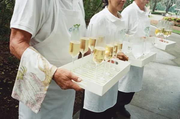 Champagne service with servers in uniform holding trays of champagne flutes on an outdoor walkway by garden shrubs and trees