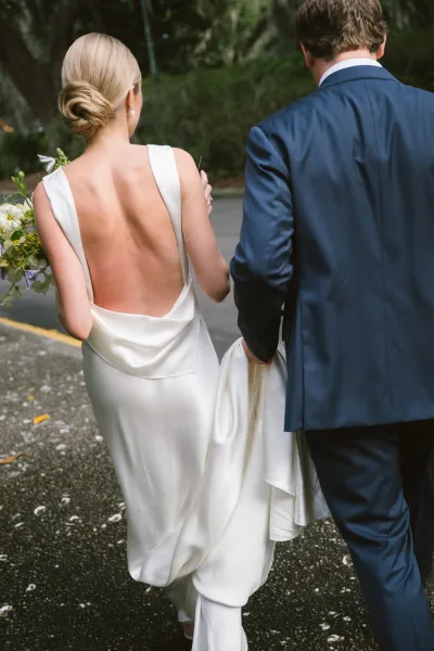 Newlywed couple holding hands, bride and groom walking away on a tree-lined path, her backless satin gown and bouquet visible