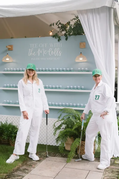 Wedding seating chart escort card display wall with golf balls on shelves, framed by ferns and string lights under a draped tent canopy