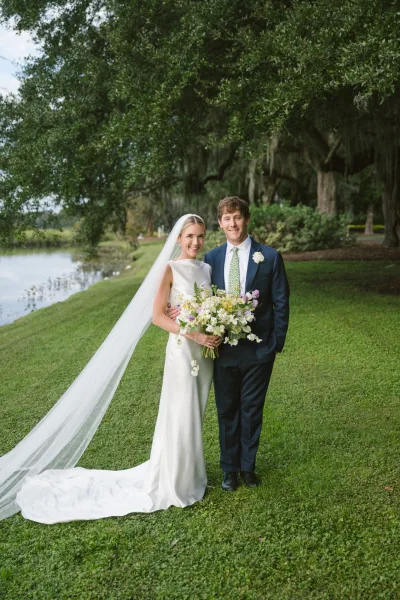 Couple portrait of bride holding bouquet beside groom in a navy suit, her long veil trailing on a lakeside lawn under mossy trees