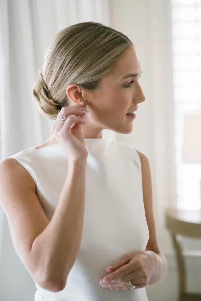 Bridal portrait of a bride getting ready, adjusting pearl stud earrings in a sleek bun, wedding dress and rings by a window with sheer curtains