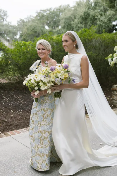 Bridal portrait of a bride holding bouquet beside her mother in a blue gown, with a long veil on a garden stone path