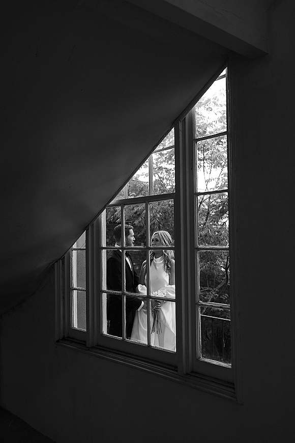 Couple portrait in a black and white wedding portrait style, bride in high-neck dress holding bouquet beside groom in tux by window