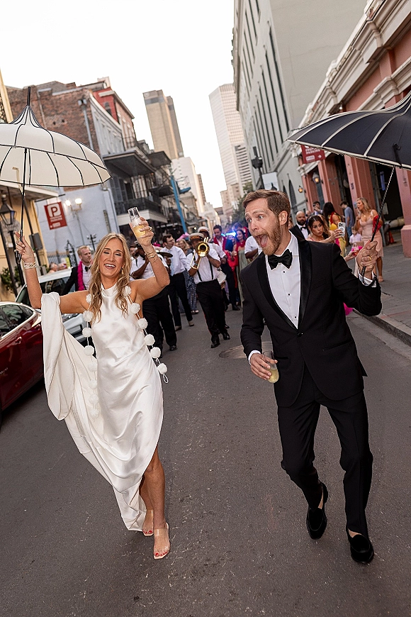 Wedding second line parade with bride and groom leading guests down a city street, holding umbrellas and drinks among historic buildings