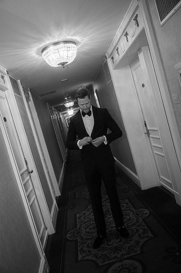Groom portrait in a black tuxedo adjusting his jacket with cufflinks, standing in a hotel hallway beneath a chandelier and doors