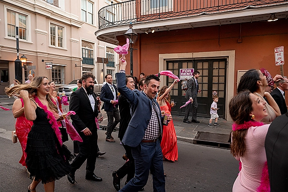 Wedding second line parade with guests in suits and cocktail dresses dancing down a city street, waving pink feather boas and bandanas