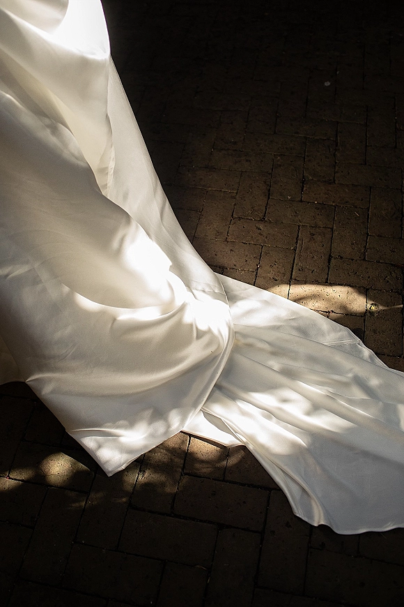 Wedding dress detail in satin fabric, the train draped on brick pavement with dappled sunlight and soft shadow patterns