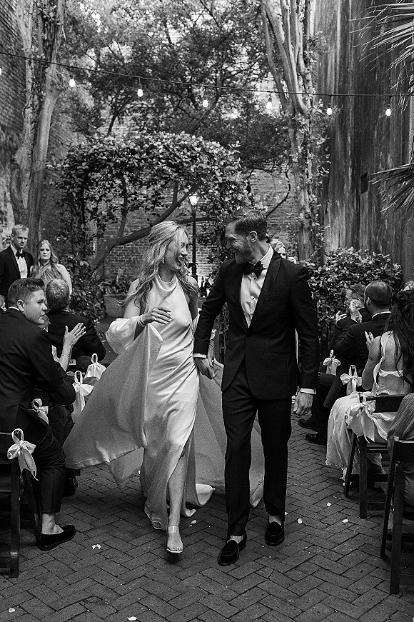 Recessional moment as newlyweds walk down the aisle under string lights, veil flowing beside tuxedoed groom in a brick courtyard
