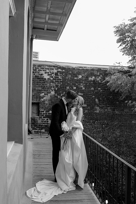 Wedding kiss portrait in a black and white wedding photo as bride in strapless gown holds bouquet while kissing groom on walkway by brick wall