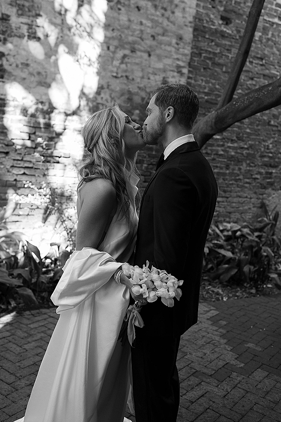 Wedding kiss portrait in black and white of bride and groom kissing in side profile, bouquet behind her back by a brick wall courtyard
