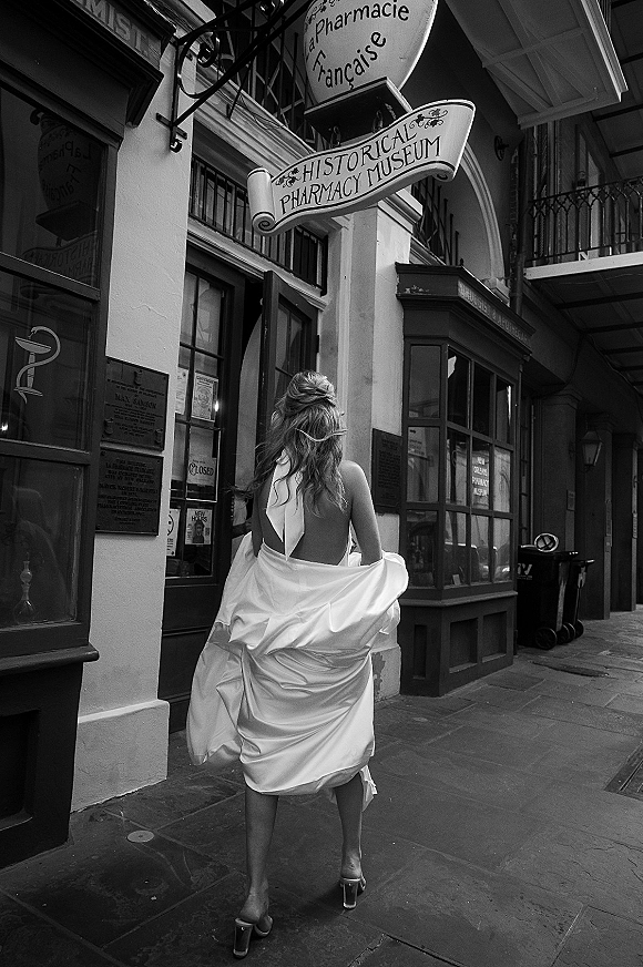 Bridal portrait of a bride walking away in an open-back wedding dress, lifting her train on a historic storefront sidewalk with awning and sign