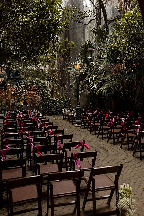 Ceremony setup with outdoor ceremony chairs, wood folding seats with pink bows under string lights in a brick courtyard with palms