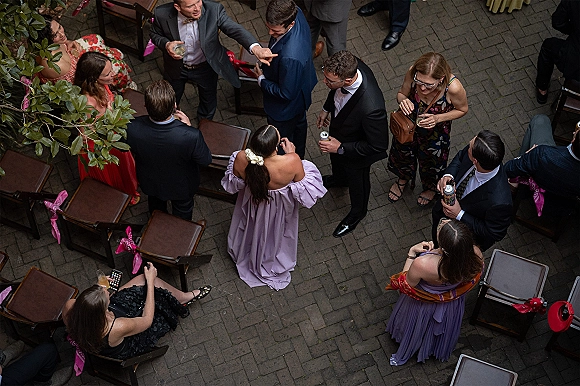 Wedding cocktail hour crowd of guests mingling in suits and colorful dresses, overhead on a brick patio with pink chair ribbons